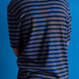 A man viewed from behind wearing a navy blue and black striped short-sleeve t-shirt, brown pants, and black shoes, standing against a solid blue studio backdrop.