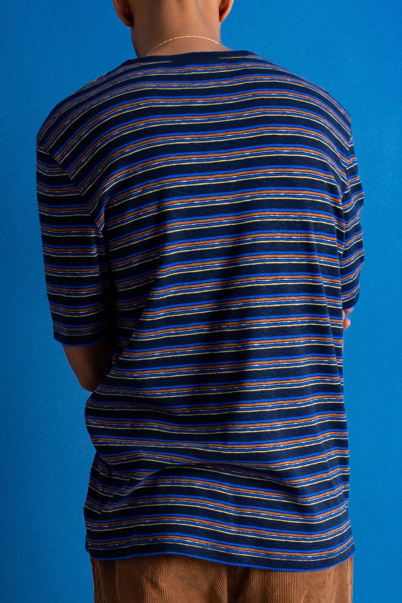 A man viewed from behind wearing a navy blue and black striped short-sleeve t-shirt, brown pants, and black shoes, standing against a solid blue studio backdrop.