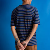 A man viewed from behind wearing a navy blue and black striped short-sleeve t-shirt, brown pants, and black shoes, standing against a solid blue studio backdrop.