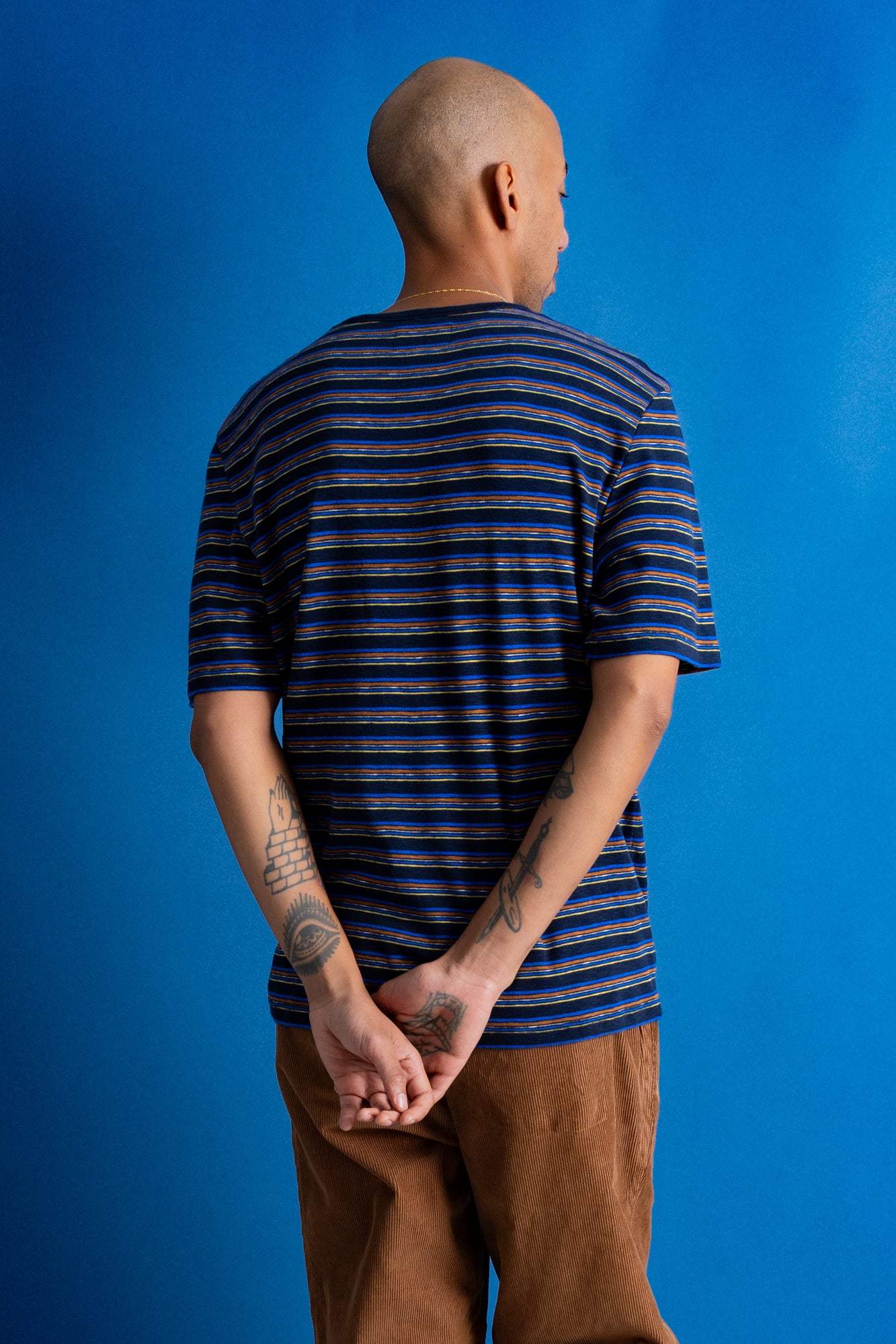 A man viewed from behind wearing a navy blue and black striped short-sleeve t-shirt, brown pants, and black shoes, standing against a solid blue studio backdrop.