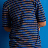 A man wearing a short-sleeve crewneck t-shirt with horizontal navy, blue, and orange stripes, paired with brown pants, set against a solid blue background.