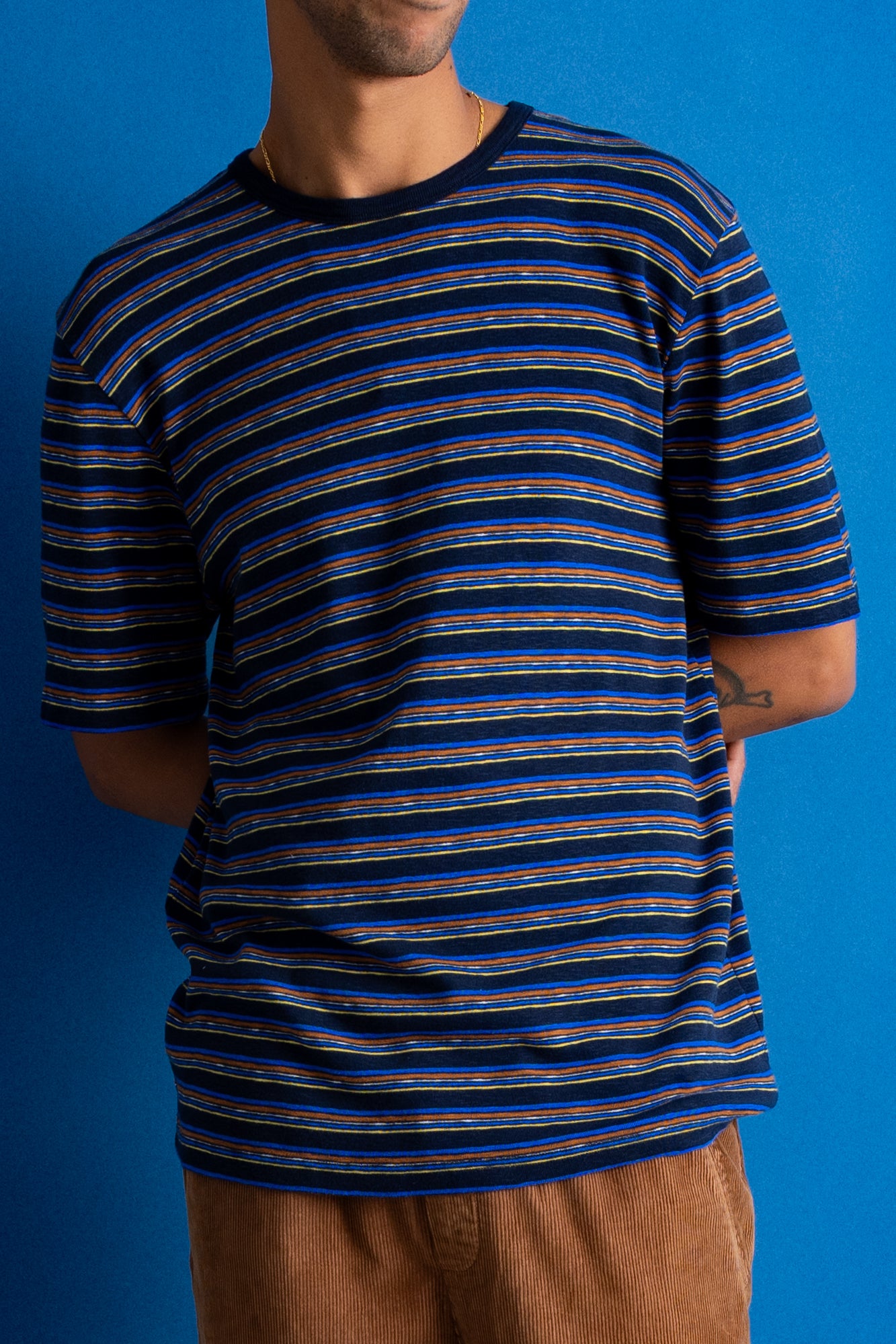 A man wearing a short-sleeve crewneck t-shirt with horizontal navy, blue, and orange stripes, paired with brown pants, set against a solid blue background.