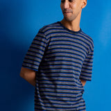 A man wearing a short-sleeve crewneck t-shirt with horizontal navy, blue, and orange stripes, paired with brown pants, set against a solid blue background.
