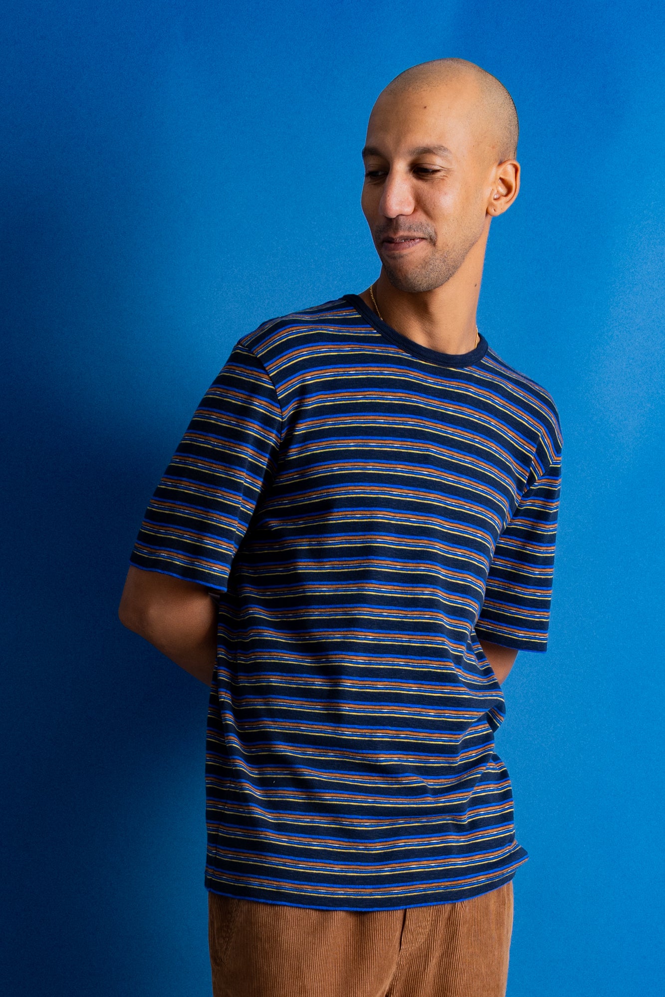 A man wearing a short-sleeve crewneck t-shirt with horizontal navy, blue, and orange stripes, paired with brown pants, set against a solid blue background.