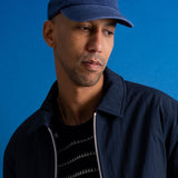 A man wearing a navy blue baseball cap with a white embroidered logo and a navy jacket over a black striped shirt, set against a solid blue background.