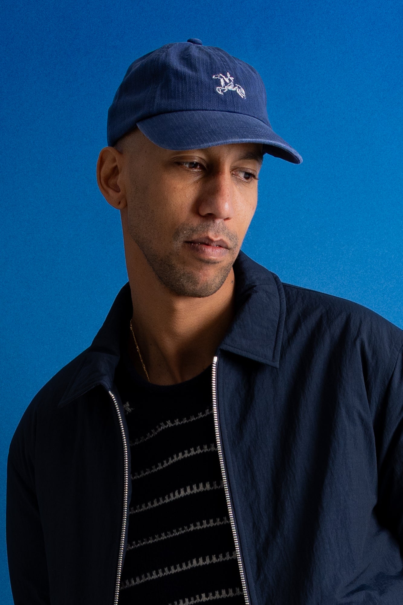 A man wearing a navy blue baseball cap with a white embroidered logo and a navy jacket over a black striped shirt, set against a solid blue background.
