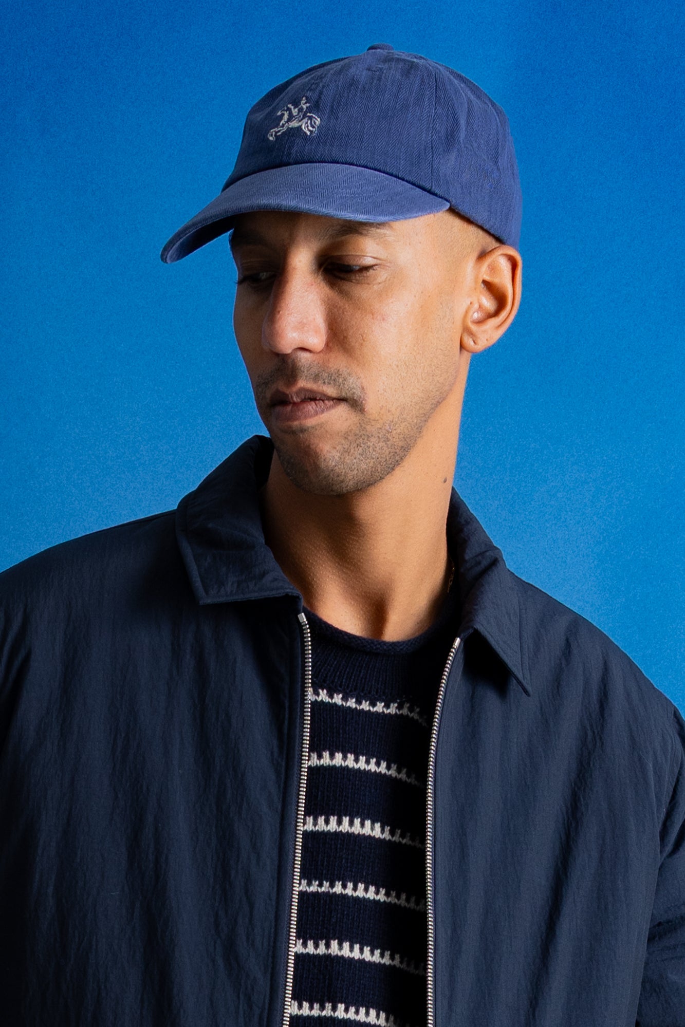 A man wearing a navy blue baseball cap with a small embroidered logo and a matching navy blue jacket over a striped knit shirt, set against a solid blue background.