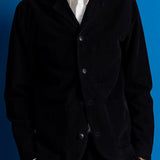 A man wearing a black corduroy blazer over a white collared shirt, standing against a solid blue background.