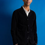 A man wearing a black corduroy blazer over a white collared shirt, standing against a solid blue background.