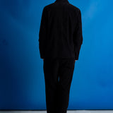 A bald man viewed from behind wearing a black corduroy blazer and matching black trousers, standing against a vibrant blue studio backdrop.