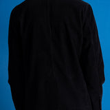 A person viewed from the back wearing a black jacket over a white collared shirt, standing against a solid blue studio background.