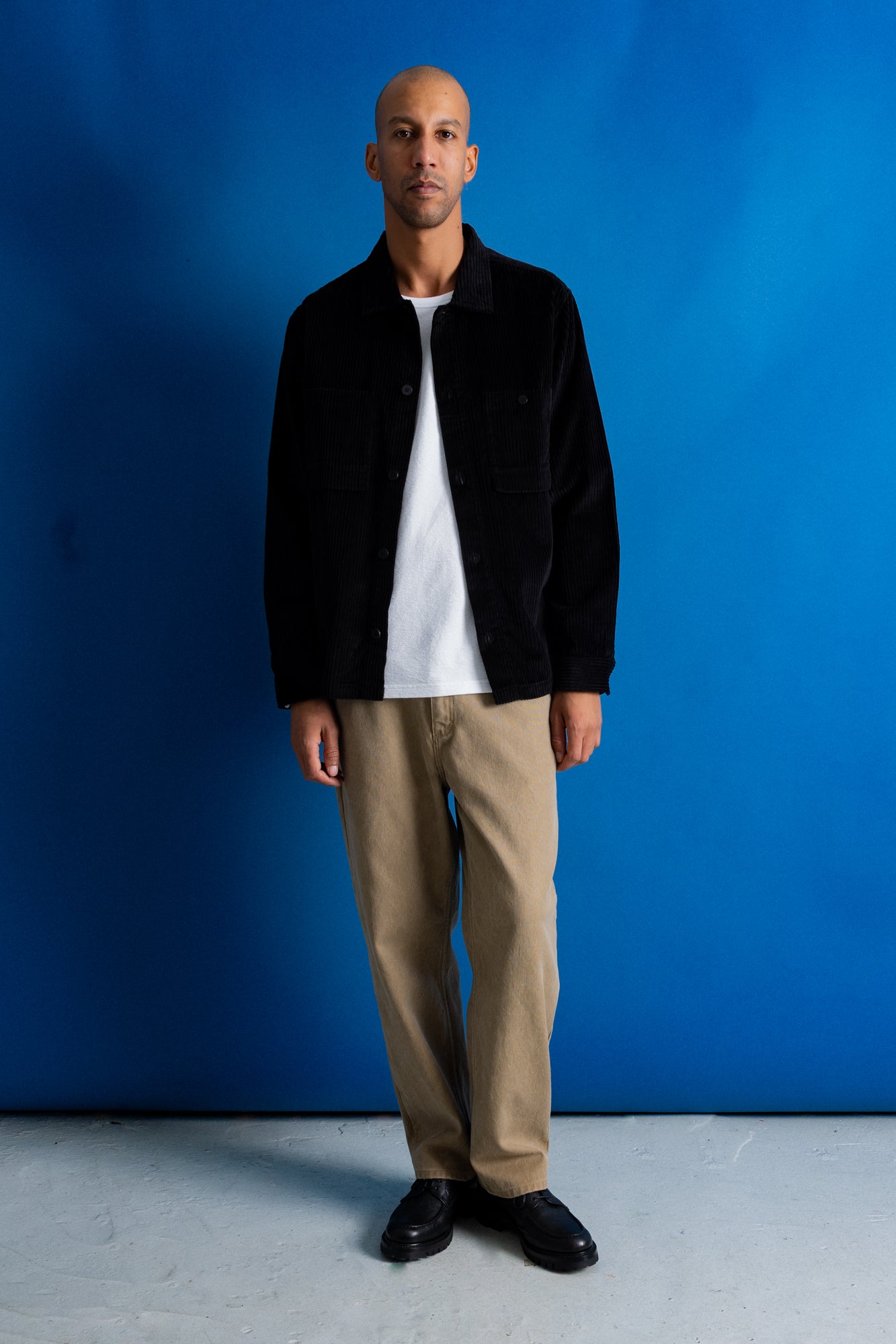 A man stands against a bright blue studio backdrop wearing a black overshirt over a white t-shirt, tan chinos, and black shoes.