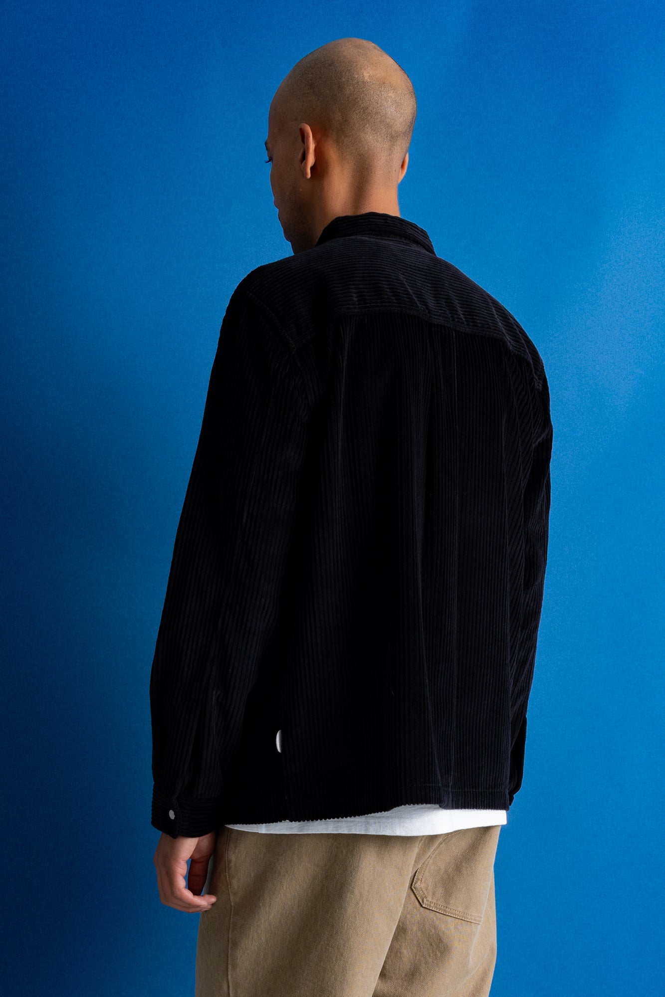 A man viewed from behind wearing a black corduroy jacket, beige trousers, and a white undershirt, set against a solid blue studio background.