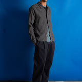 A man wearing a dark gray textured shirt, black trousers, a light blue t-shirt, and a dark cap stands against a vibrant blue studio backdrop.