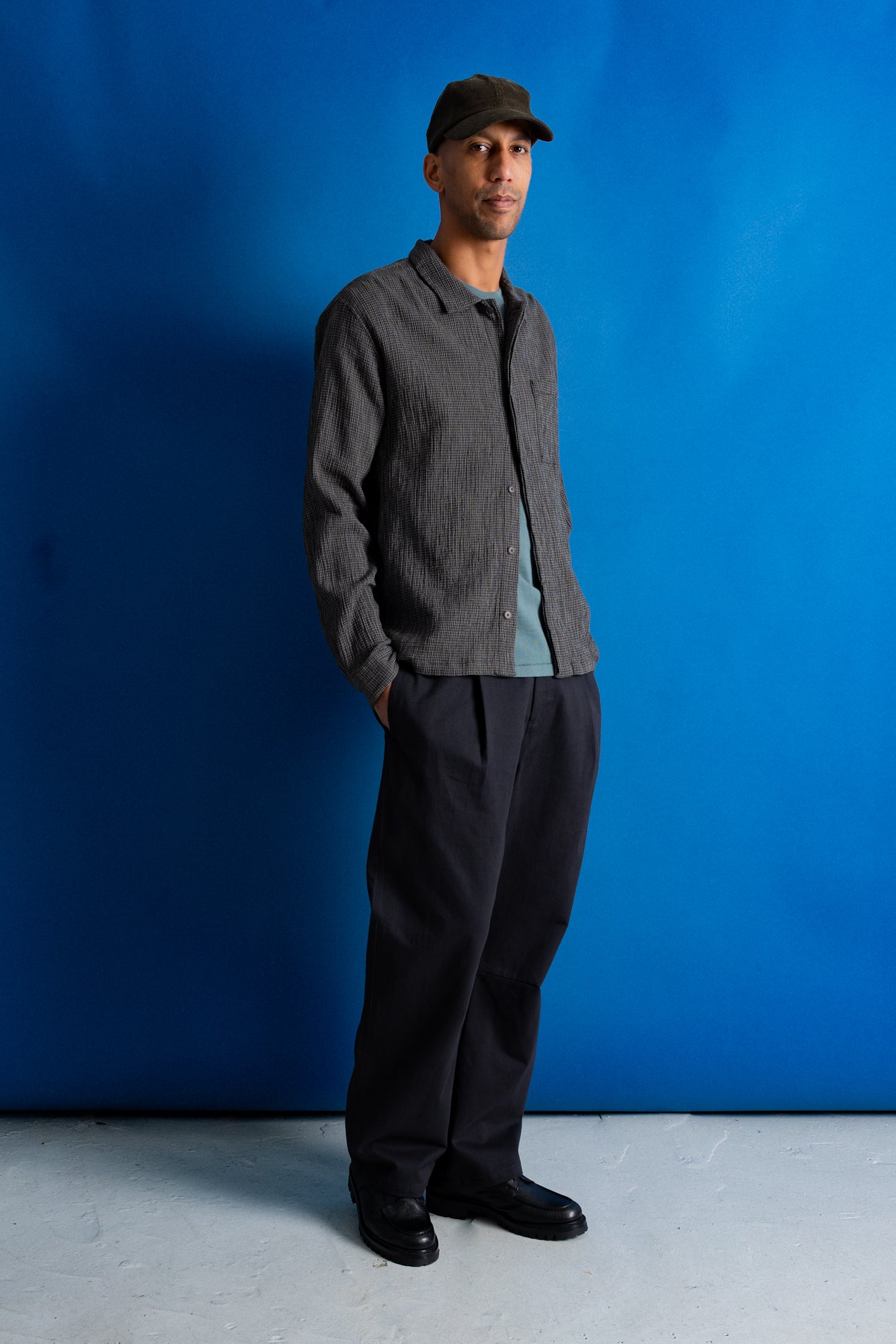 A man wearing a dark gray textured shirt, black trousers, a light blue t-shirt, and a dark cap stands against a vibrant blue studio backdrop.