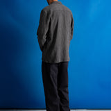 Man facing away from the camera wearing textured grey checkered shirt, dark grey trousers and a dark green cap against a bright blue background