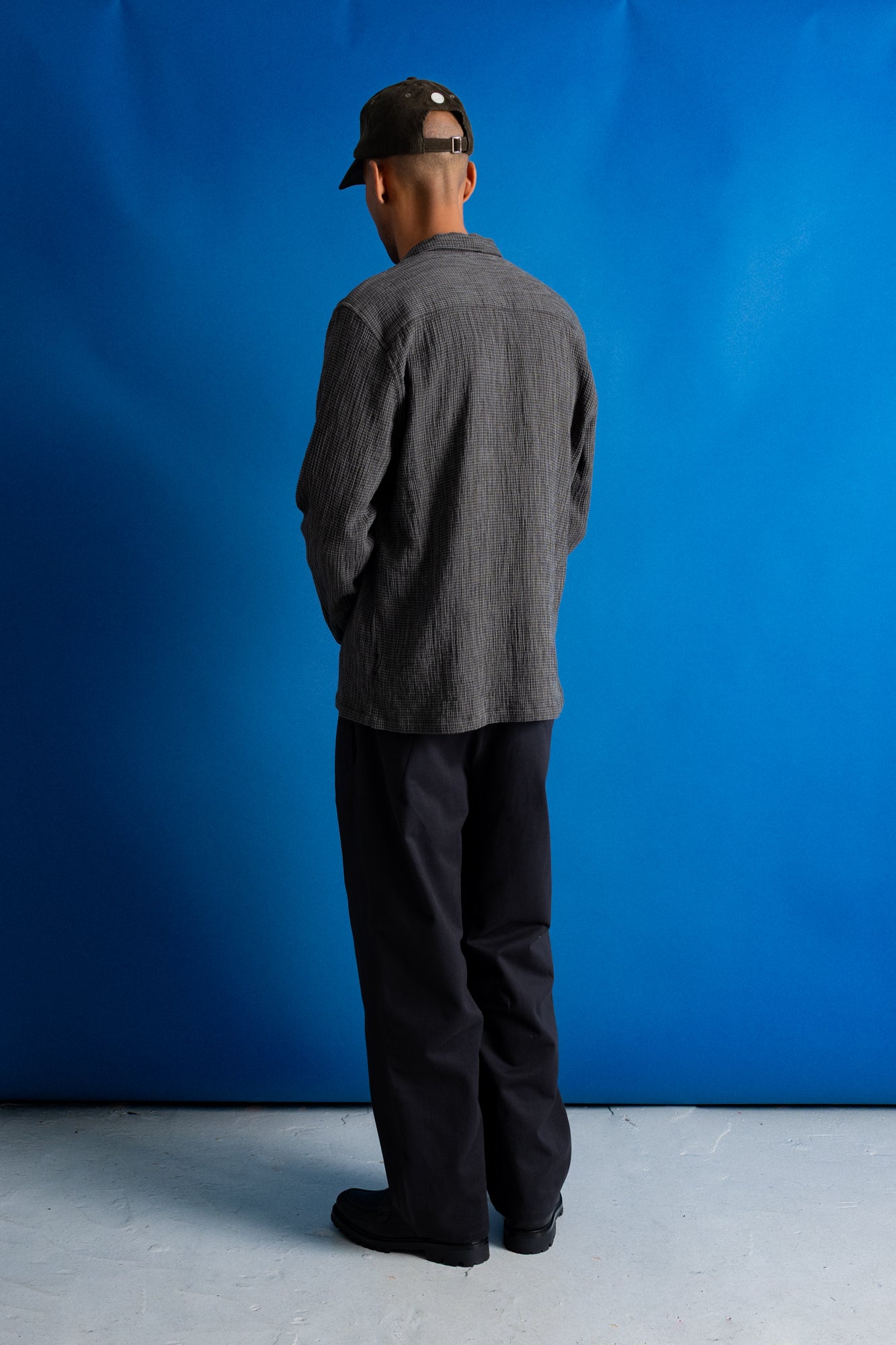 Man facing away from the camera wearing textured grey checkered shirt, dark grey trousers and a dark green cap against a bright blue background