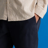 Close-up of a person wearing a light beige linen button-up shirt and dark navy trousers, standing against a solid blue background.