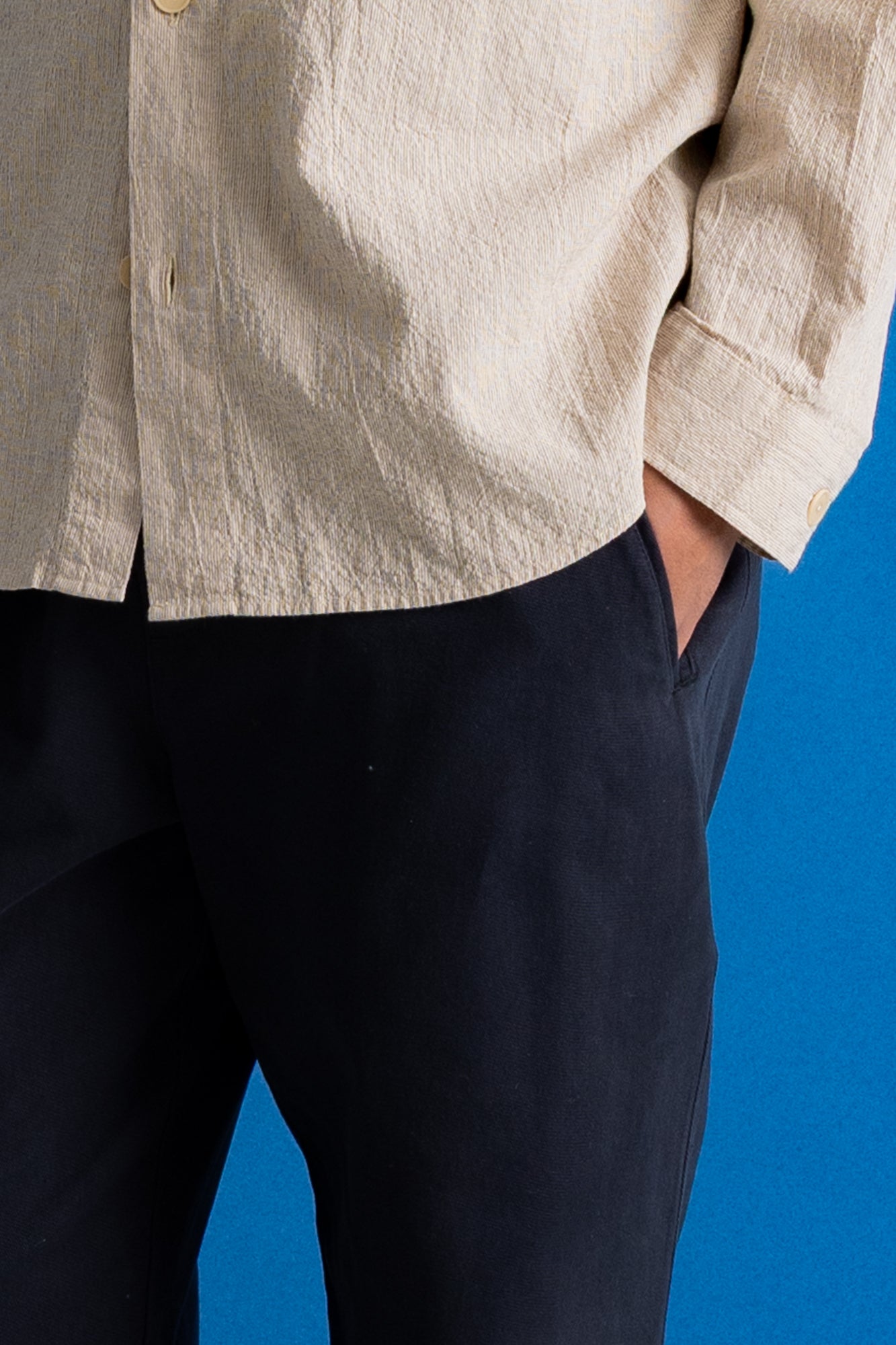 Close-up of a person wearing a light beige linen button-up shirt and dark navy trousers, standing against a solid blue background.