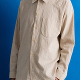 Close-up of a person wearing a light beige linen button-up shirt and dark navy trousers, standing against a solid blue background.