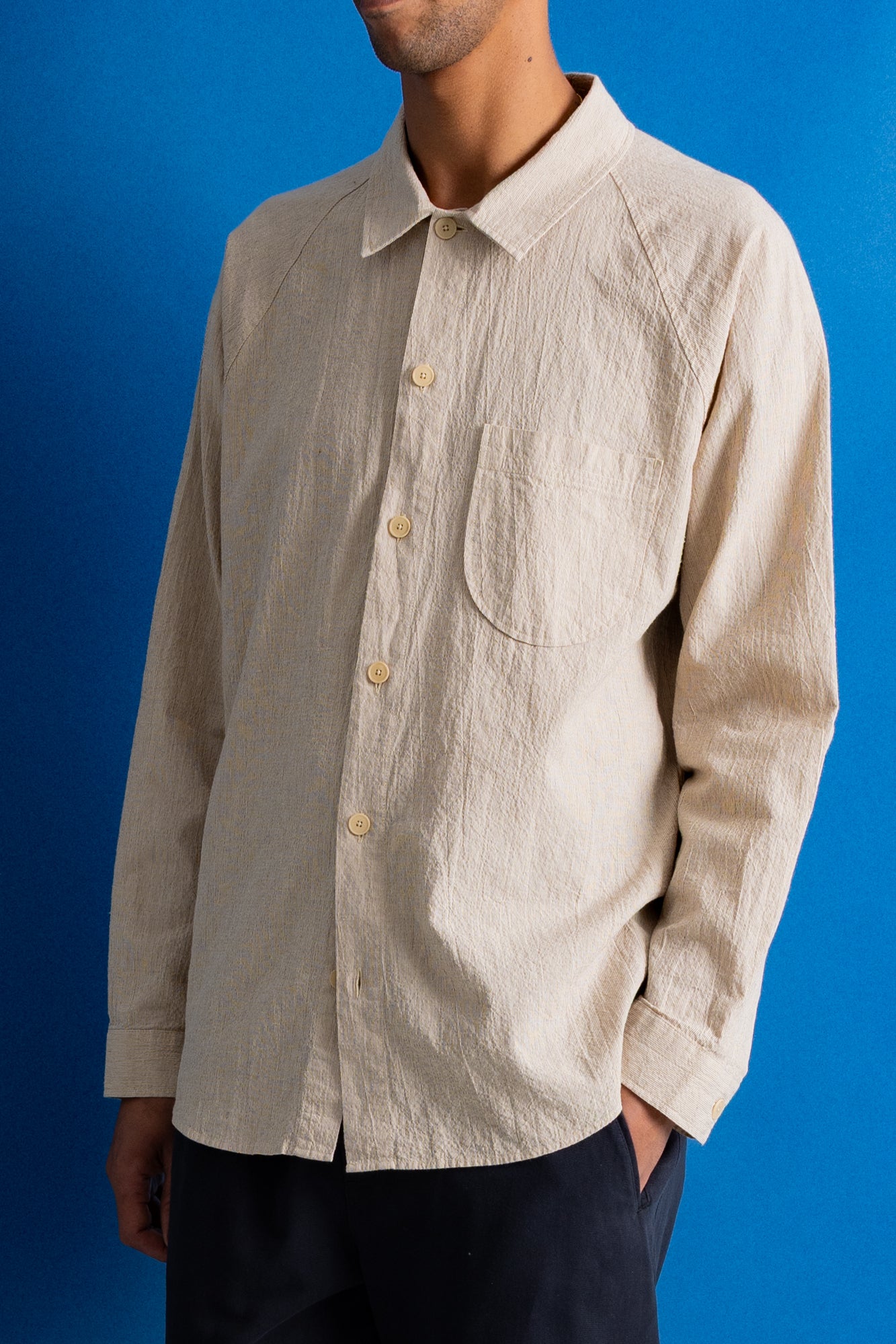 Close-up of a person wearing a light beige linen button-up shirt and dark navy trousers, standing against a solid blue background.