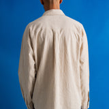 A man viewed from behind wearing a light beige textured long-sleeve button-up shirt and dark trousers, standing against a solid blue background.
