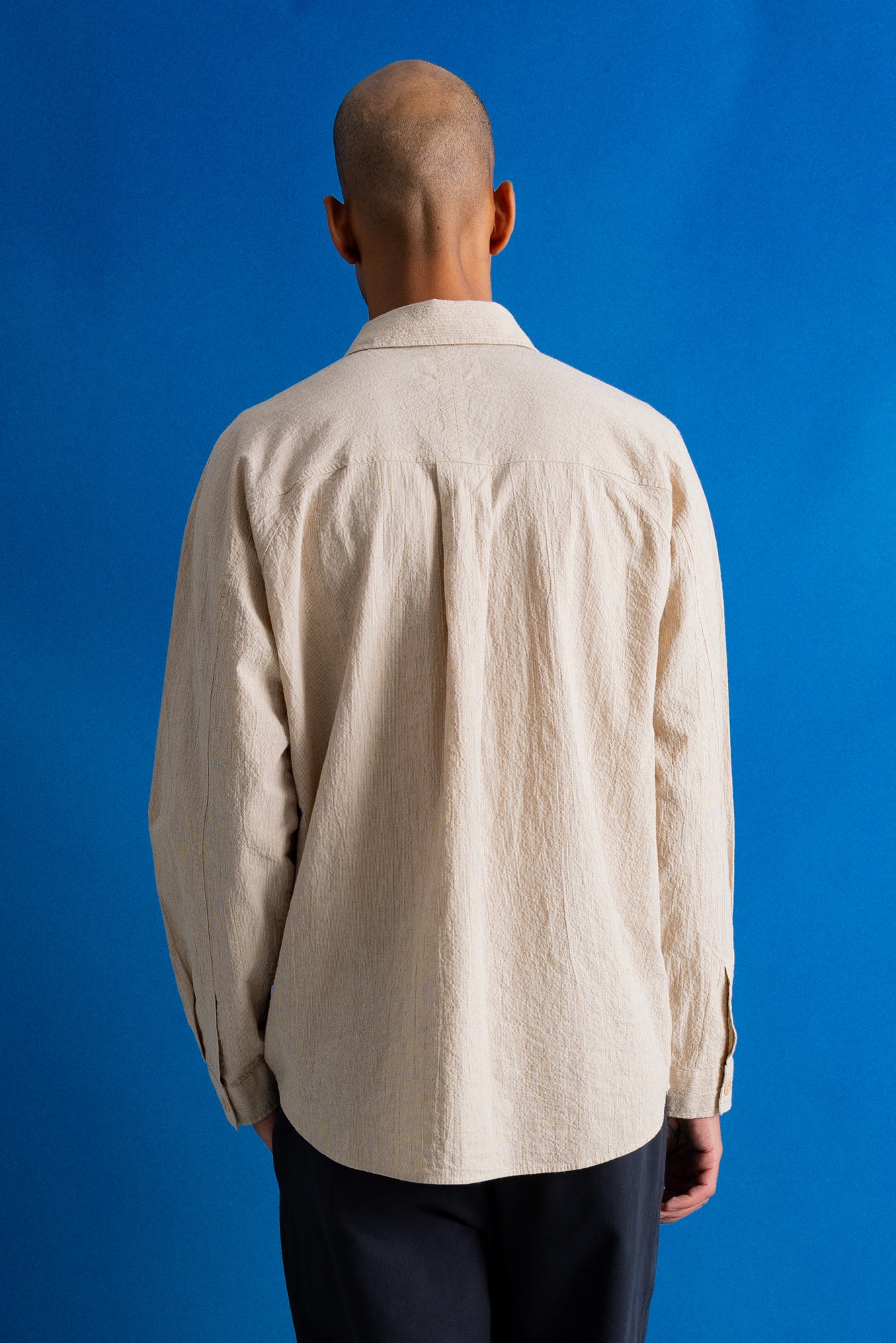 A man viewed from behind wearing a light beige textured long-sleeve button-up shirt and dark trousers, standing against a solid blue background.