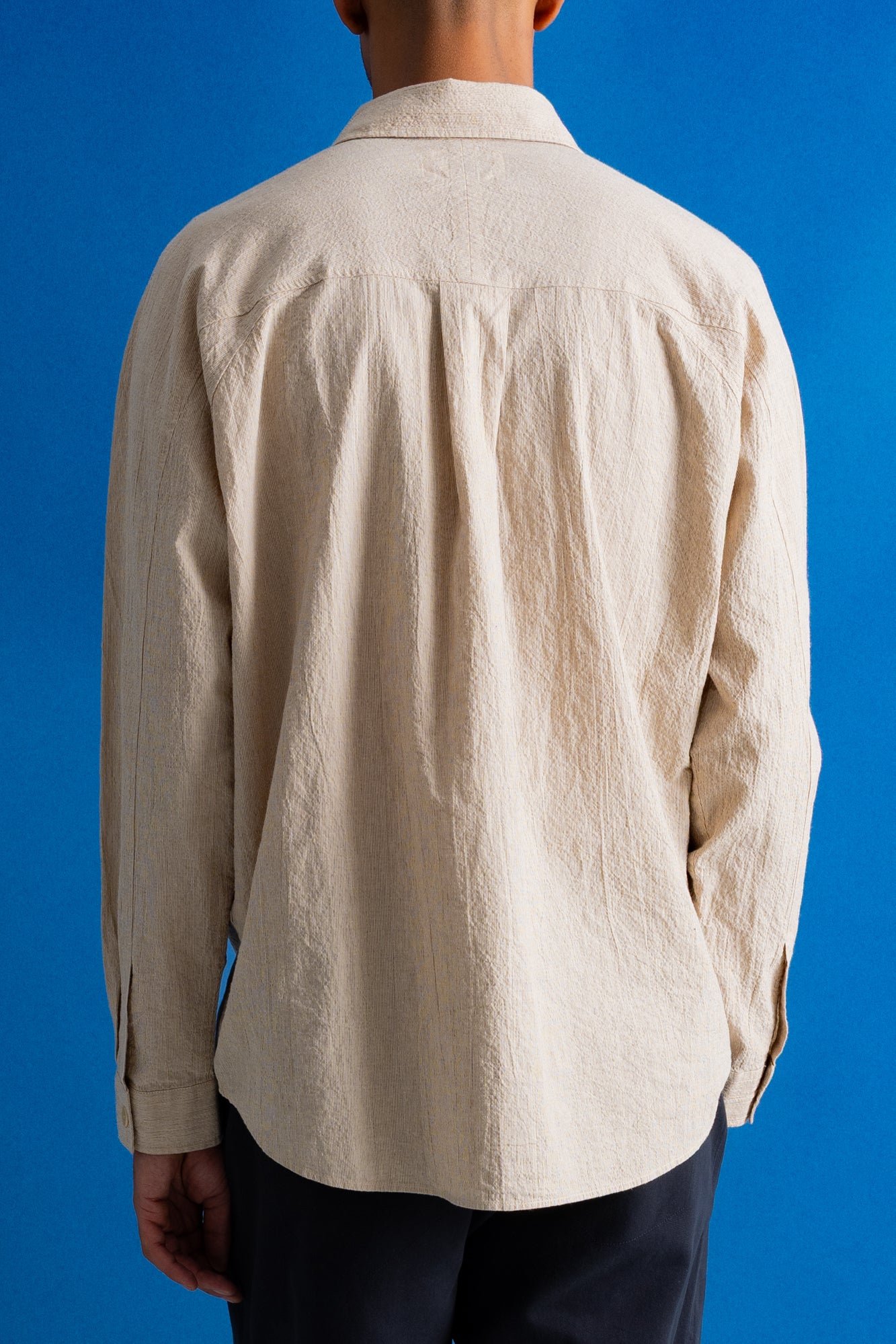 A person viewed from the back wearing a light beige linen button-up shirt and dark trousers, set against a solid blue background.
