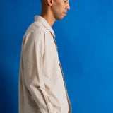 A man in profile wearing a light beige textured linen button-down shirt and dark trousers, standing against a solid blue studio background.
