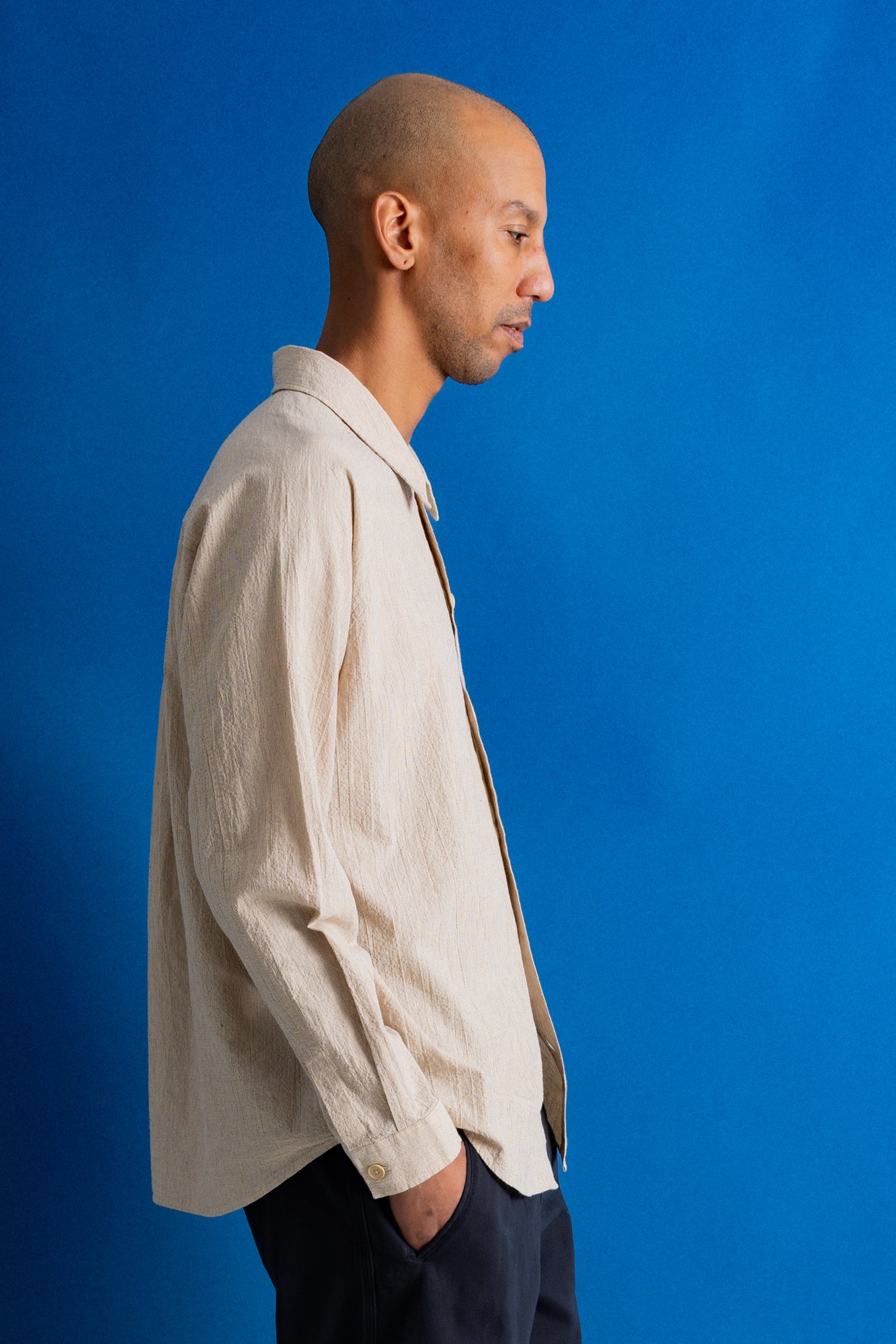 A man in profile wearing a light beige textured linen button-down shirt and dark trousers, standing against a solid blue studio background.