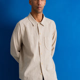 A man wearing a light beige textured long-sleeve button-up shirt and dark pants, standing against a solid blue background.