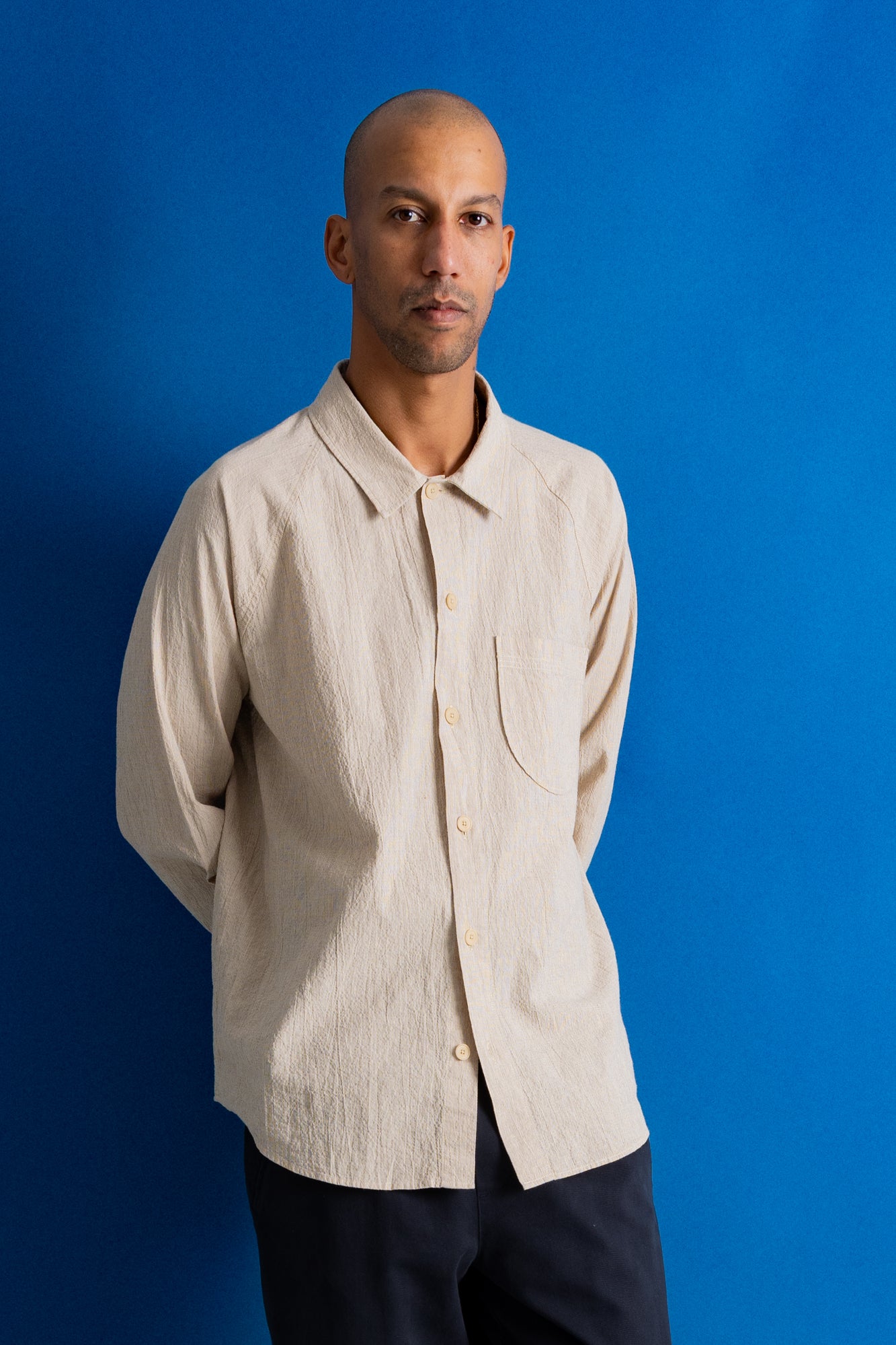 A man wearing a light beige textured long-sleeve button-up shirt and dark pants, standing against a solid blue background.