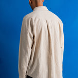 A man viewed from behind wearing a light beige textured linen shirt and dark trousers, standing against a solid blue background.