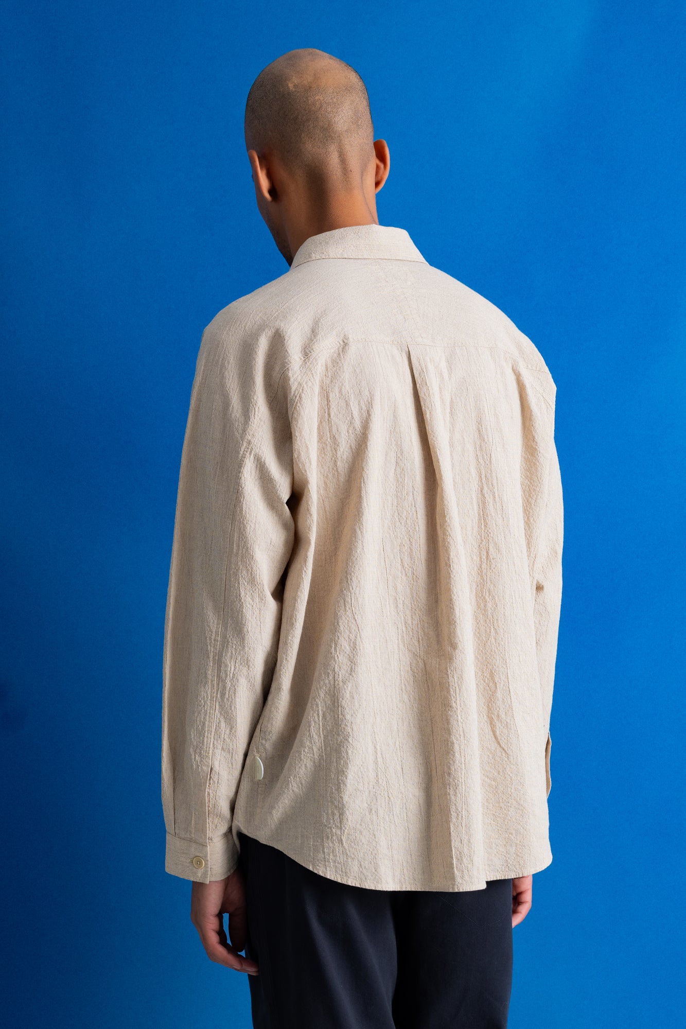 A man viewed from behind wearing a light beige textured linen shirt and dark trousers, standing against a solid blue background.