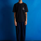 A man stands against a bright blue studio backdrop wearing a black short-sleeve T-shirt with a small white chest graphic and loose dark pants.