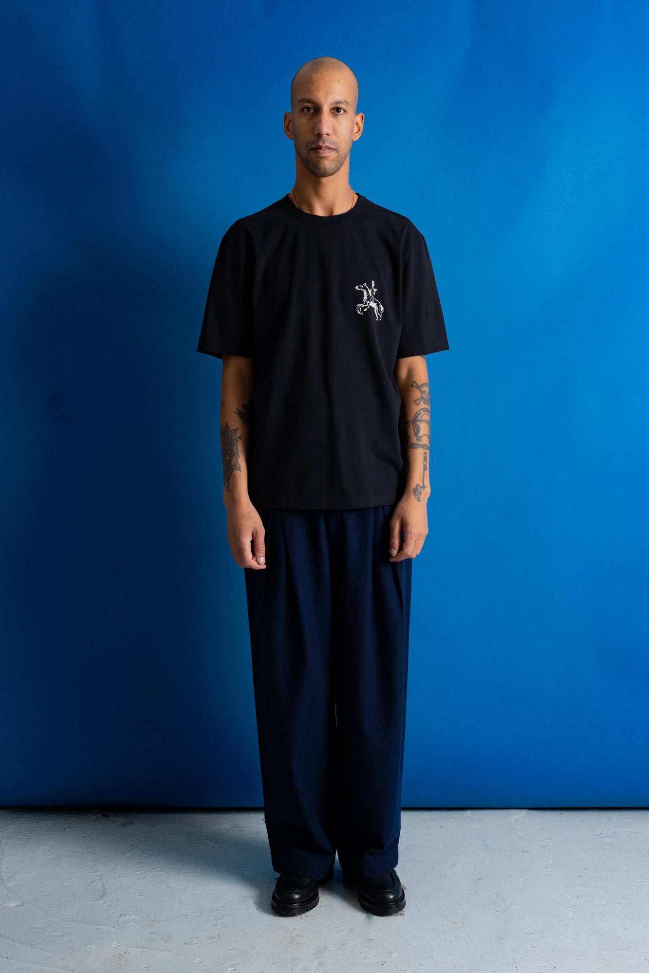 A man stands against a bright blue studio backdrop wearing a black short-sleeve T-shirt with a small white chest graphic and loose dark pants.