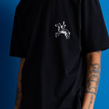 A man wearing a black short-sleeve t-shirt with a white graphic of a knight on a rearing horse on the left chest, posed against a solid blue background.