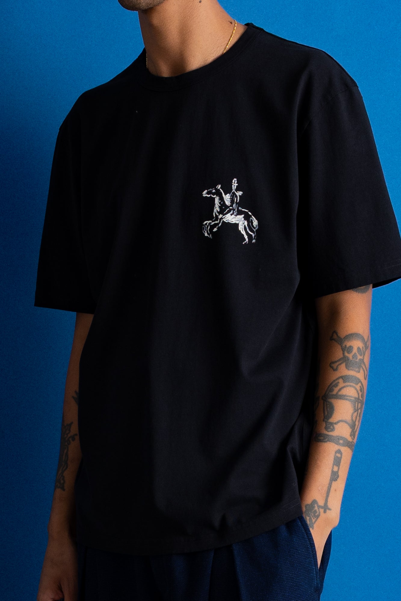 A man wearing a black short-sleeve t-shirt with a white graphic of a knight on a rearing horse on the left chest, posed against a solid blue background.