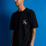 A man wearing a black short-sleeve t-shirt with a small white graphic of a knight on a horse on the left chest, posed against a solid blue background.