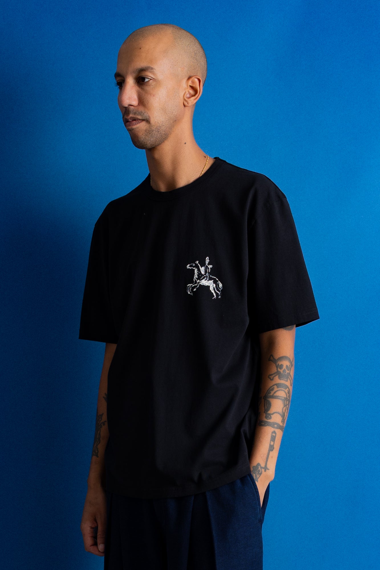 A man wearing a black short-sleeve t-shirt with a small white graphic of a knight on a horse on the left chest, posed against a solid blue background.