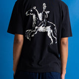 A man facing away from the camera, wearing a black short-sleeve t-shirt with a white graphic of a knight on the back, posed against a solid blue background.