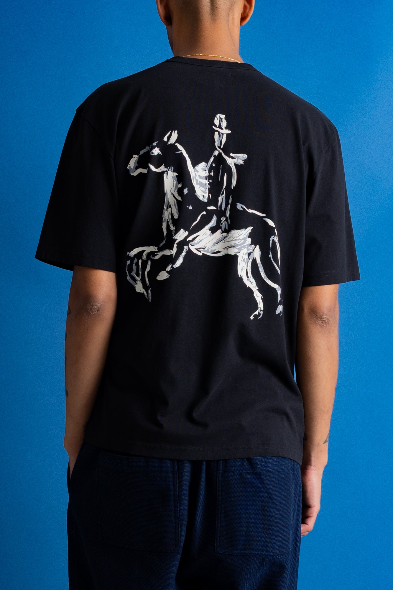 A man facing away from the camera, wearing a black short-sleeve t-shirt with a white graphic of a knight on the back, posed against a solid blue background.