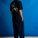 A man facing away from the camera, wearing a black short-sleeve t-shirt with a white graphic of a knight on the back, posed against a solid blue background.