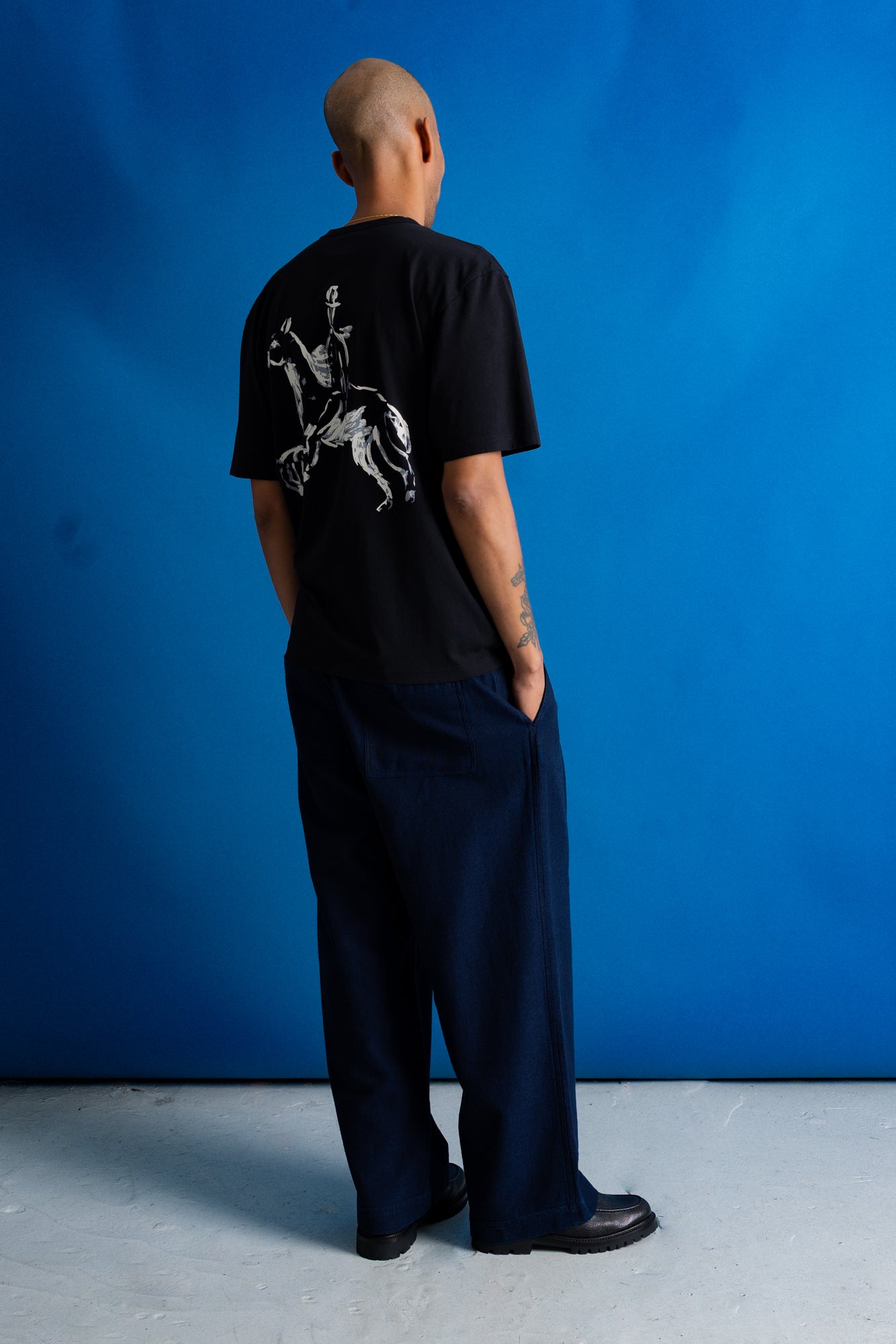 A man facing away from the camera, wearing a black short-sleeve t-shirt with a white graphic of a knight on the back, posed against a solid blue background.