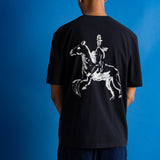 A man viewed from behind wearing a black t-shirt with a large white graphic of a rider on a horse across the back, set against a solid blue background.