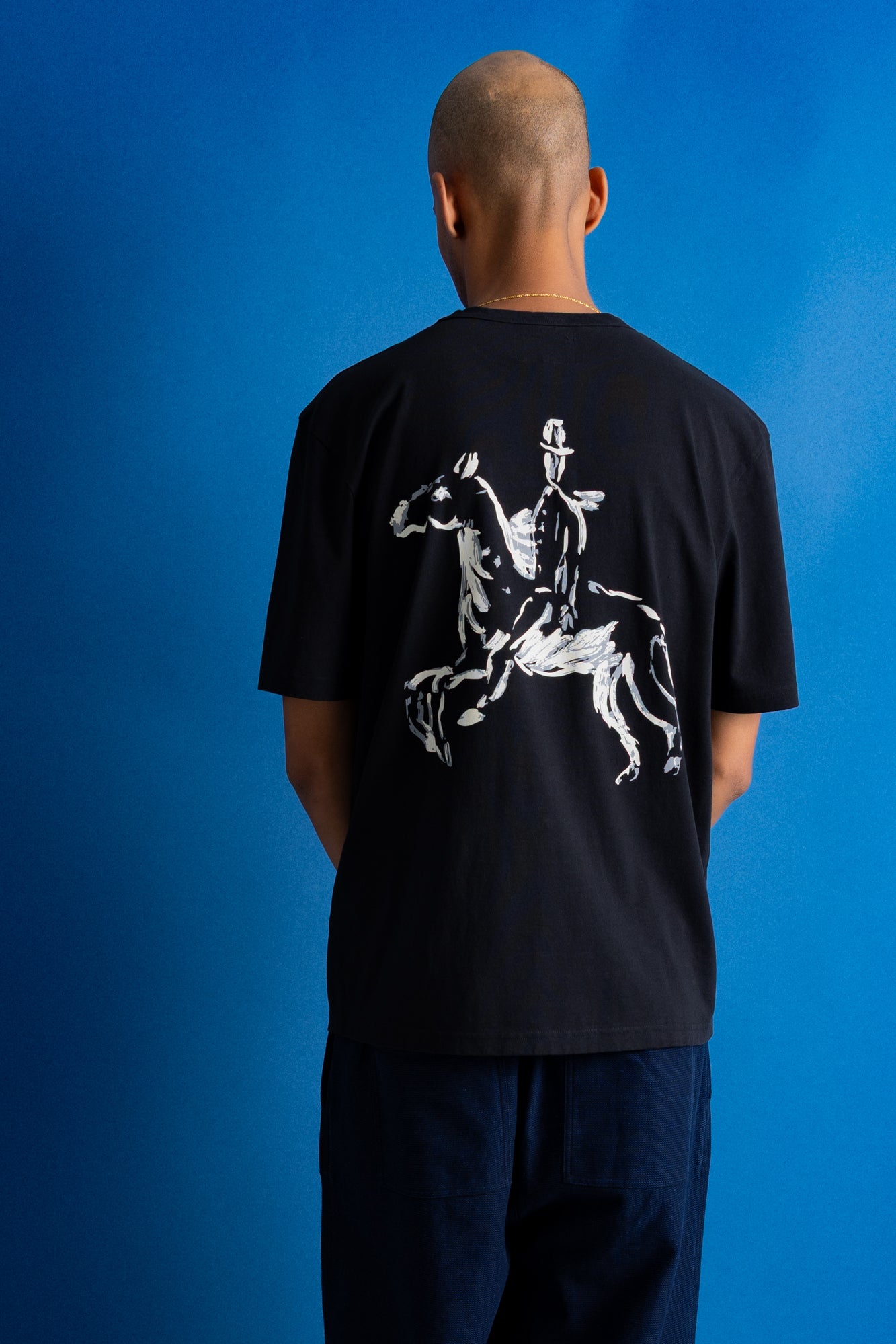 A man viewed from behind wearing a black t-shirt with a large white graphic of a rider on a horse across the back, set against a solid blue background.