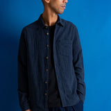 A man wearing a dark navy blue textured button-up shirt over a black t-shirt and navy pants, standing against a solid blue studio background.