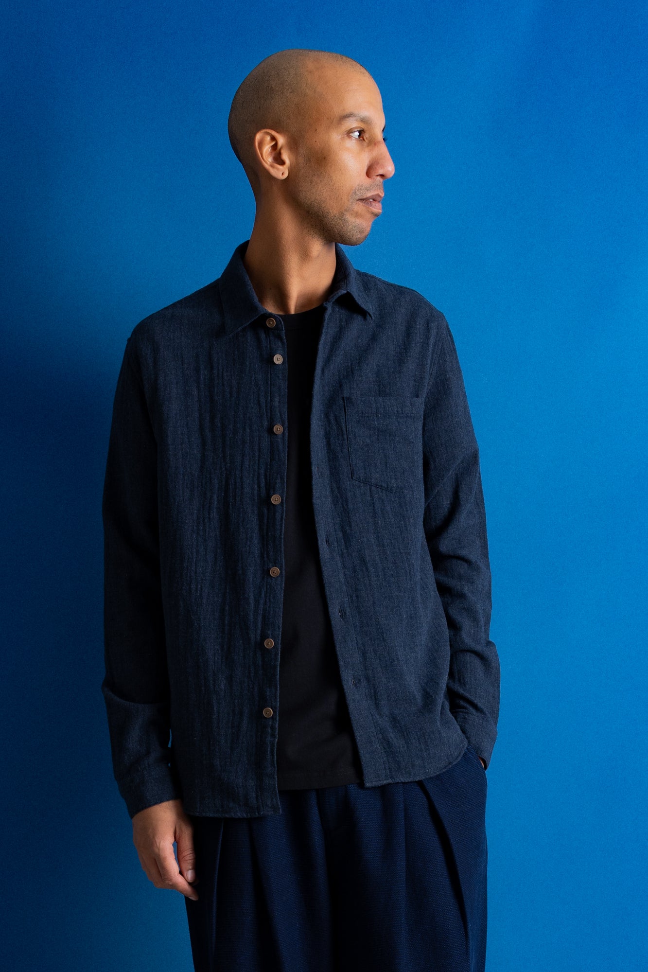 A man wearing a dark navy blue textured button-up shirt over a black t-shirt and navy pants, standing against a solid blue studio background.