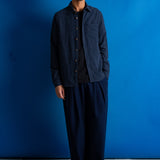 A man stands against a vibrant blue studio backdrop wearing a dark navy blue button-up shirt with vertical pleats over a black t-shirt, paired with matching dark navy pleated trousers and black shoes.