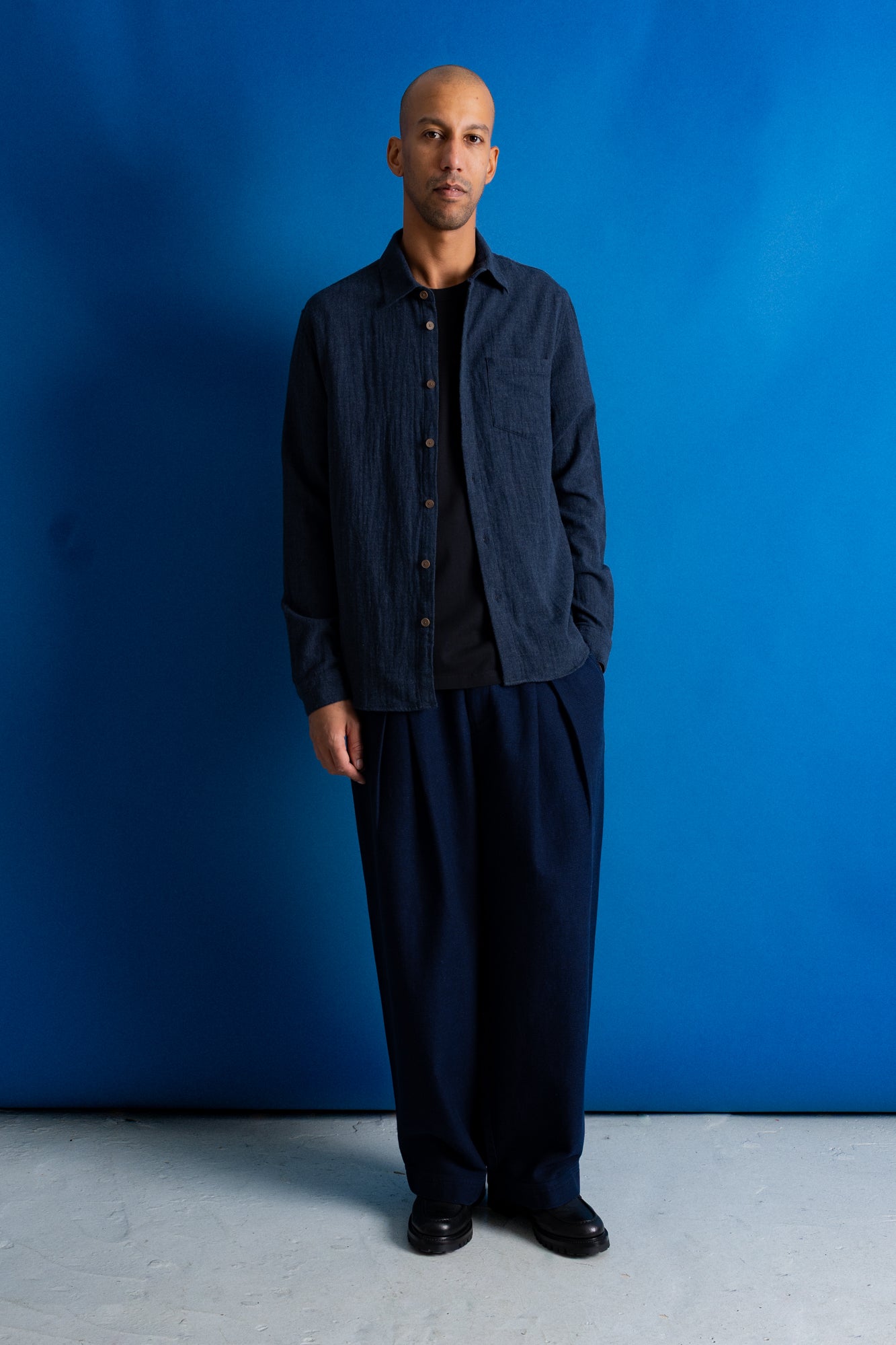 A man stands against a vibrant blue studio backdrop wearing a dark navy blue button-up shirt with vertical pleats over a black t-shirt, paired with matching dark navy pleated trousers and black shoes.